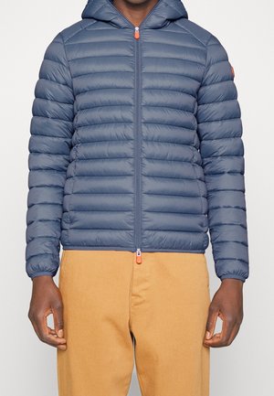 Marineblaue Steppjacke mit horizontalem Rautenmuster, Kapuze und Frontreißverschluss. Mit orangefarbenen Reißverschlussakzenten und elastischen Bündchen. Kombiniert mit beigen Hosen.