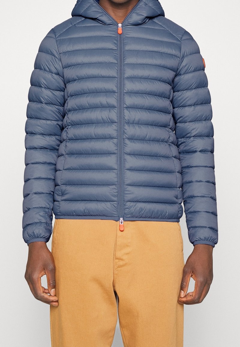 Navy puffer jacket with horizontal quilted design, hood, and front zipper. Features orange zipper accents and elastic cuffs. Paired with tan pants.