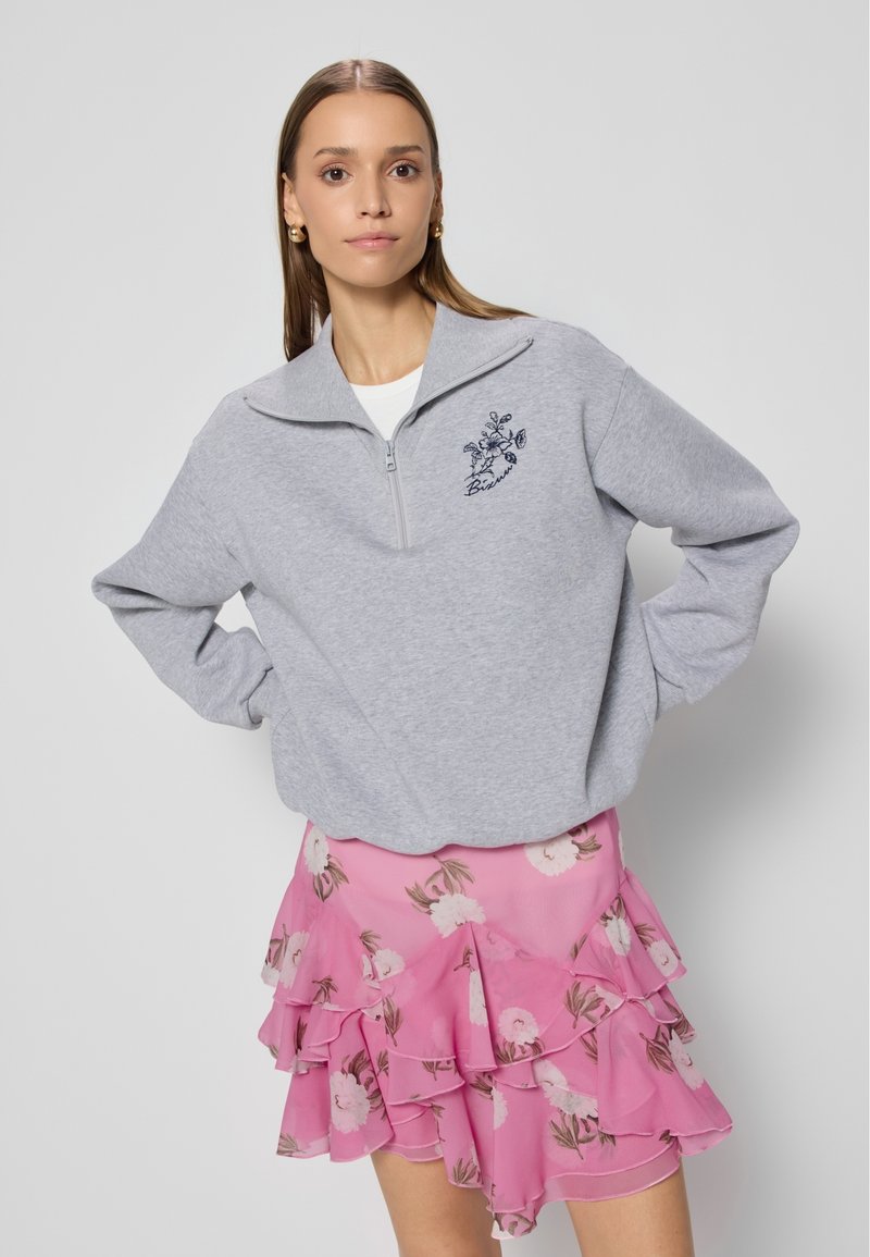 Grijze rits sweatshirt met geborduurd bloemenpatroon op de borst, gecombineerd met een roze gerimpelde rok met een bloemenprint in wit en groen.