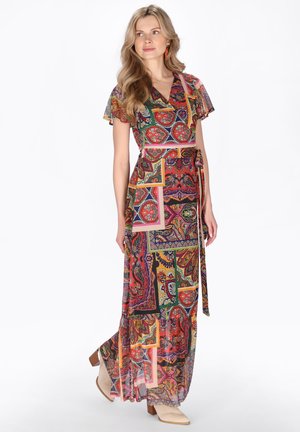 Vrouw met lang blond haar, gekleed in een kleurrijke maxi-jurk met patroon en beige enkellaarzen, staande tegen een witte achtergrond.