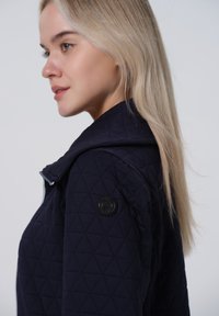 Veste matelassée bleu marine avec col haut, motif en triangle texturé, fermeture éclair et un patch rond noir sur l'épaule gauche.