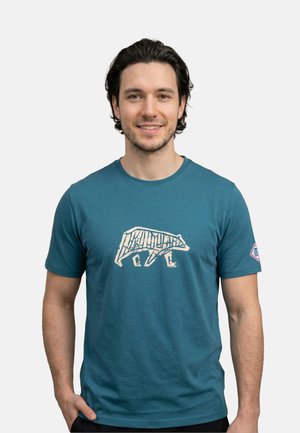 Jonge man met donker haar die een turquoise T-shirt met een witte abstracte berenafbeelding draagt, staand tegen een effen lichte achtergrond.