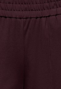 Burgunderfarget stoff med en jevn tekstur og elastisk midjebånd. Materialet er lett med synlige sømdetaljer langs sømmene.