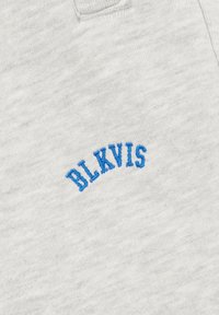 Tela gris claro con texto bordado en azul "BLKVIS" en una fuente de bloque curvada cerca del borde de un bolsillo.