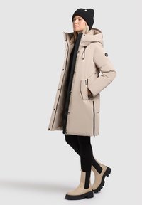 Manteau matelassé beige avec capuche, boutons devant et poches zippées. Porté par-dessus une tenue noire et associé à des bottines chunky beige.