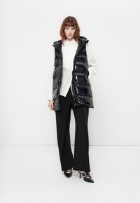 Vrouw in een glanzend zwart puffervest, witte blouse, zwarte wijde broek en zwarte puntige hakken met banden, staand tegen een witte muur.