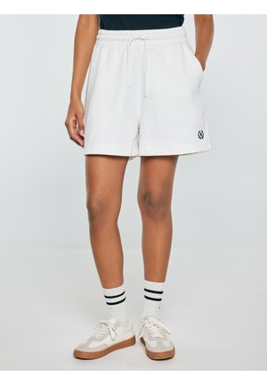 Osoba nosząca białe spodenki ze ściągaczem i bocznymi kieszeniami, białe skarpetki z dwoma czarnymi paskami oraz białe trampki z beżowymi podeszwami.