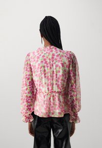 Blouse fleurie avec motifs roses et verts, tissu transparent, manches longues bouffantes, détail à la taille froncée, associée à un pantalon noir brillant.