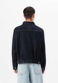 Donkerdenim jasje gezien van achteren, met een kraag, knoopaccenten en een iets losse pasvorm. Eenvoudig ontwerp met zichtbare stiksels.