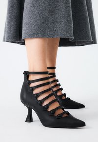 Zapatos de tacón alto negros con puntas afiladas, que presentan múltiples correas horizontales adornadas con botones circulares y un diseño de tacón triangular único.