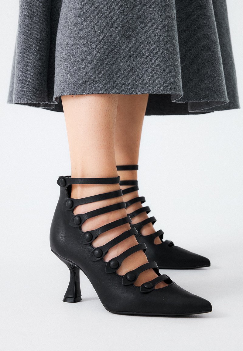 Zapatos de tacón alto negros con puntas afiladas, que presentan múltiples correas horizontales adornadas con botones circulares y un diseño de tacón triangular único.