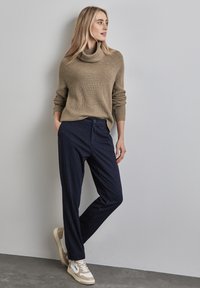 Beige grovstickad polokrage tröja i kombination med marinblå randiga byxor och beiga sneakers, lutande mot en ljusgrå vägg.