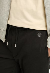 Schwarze Sweatpants mit Kordelzug, versehen mit einem dezenten grauen Logo. Getragen mit einem beigen Shirt und einem grauen Reißverschluss-Hoodie. Weiches Baumwollmischgewebe.