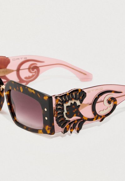 Occhiali da sole quadrati tartarugati con aste rosa che presentano intricati vortici neri e lenti scure sfumate su uno sfondo bianco.