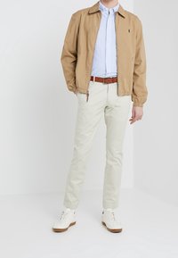 Jasnobrązowa kurtka na suwak, niebieska koszula w paski, beżowe spodnie i białe trampki z brązowymi podeszwami. Kurtka ma elastyczne mankiety i kołnierz.