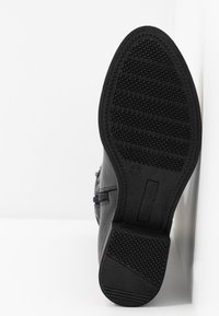 Semelle de botte noire inclinée contre une surface blanche, montrant un motif de bande de roulement avec des crêtes et des sections de grip texturées.