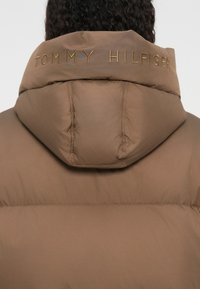 Hnědá puffová bunda s kapucí, prošívanou texturou a zlatě vyšitým logem "TOMMY HILFIGER" na zadní straně.