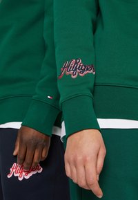 Felpa verde con logo "Hilfiger" ricamato in rosso e bianco sulla manica, abbinata a pantaloni blu navy che mostrano un logo simile.