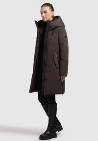 Dlhý hnedý puffový kabát s kapucňou, so zipsom na bočnej vrecku, s tlačidlovými uzávermi a výrazným prešívaním; kombinovaný s čiernymi legínami a vysokými čižmami.
