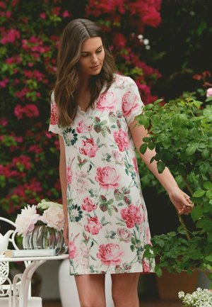 Woman in white floral dress touching green rose plant beside white table with flowers in a garden with pink blossoms background.