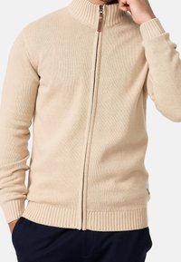 Pull beige à fermeture éclair, avec col et poignets côtelés, arborant un motif en maille texturée et un détail de tirette en bois.