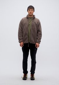 Μπουφάν από φλις σε μπεζ, με φερμουάρ σε τσέπη και ψηλό γιακά, φορεμένο πάνω από ένα πράσινο μπλουζάκι και συνδυασμένο με μαύρο παντελόνι και μποτάκια πεζοπορίας.