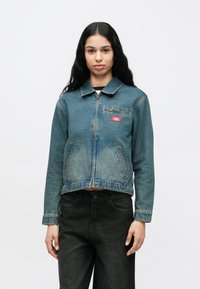 Γυναίκα με μακριά σκούρα μαλλιά που φοράει ξεθωριασμένο μπλε τζιν μπουφάν με φερμουάρ και μαύρο τζιν, στέκεται μπροστά σε απλό λευκό φόντο.