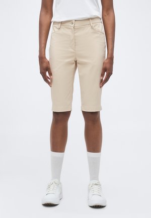 Oseba z bež kratkimi hlačami do kolen, belimi visokimi nogavicami, belimi supergami in belo srajco, stoji z rokami sproščeno ob straneh.