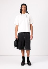 Tricou alb cu mânecă scurtă, pantaloni scurți negri din denim și cizme negre de tip combat. Modelul ține o geantă mică neagră. Design minimalist, croială relaxată, culori contrastante.