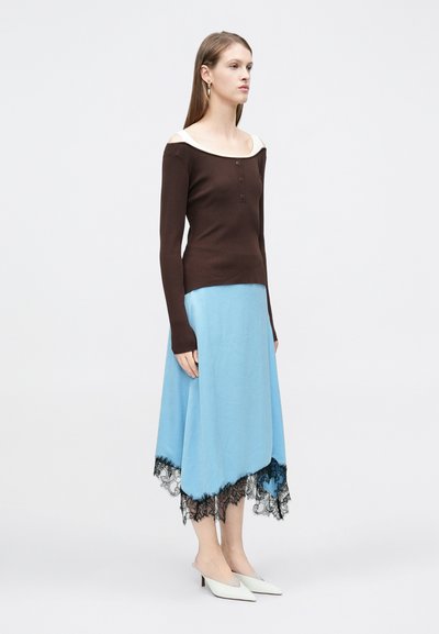 Καφέ μακρυμάνικο τοπ με ριμπ ύφανση και εκρού λαιμόκοψη εκτός ώμων, σε συνδυασμό με φούστα τιρκουάζ με μαύρο δαντελένιο τελείωμα και λευκά mule με τακούνι.