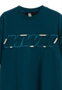 Camiseta de algodón en color azul verdoso que presenta un gran logo bordado "MWM" en negro y azul, con detalles metálicos adheridos a las letras.