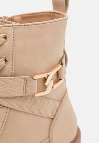 Beige syntetická kůže kotníková bota s embosovaným trojúhelníkovým vzorem, zlatým kováním a béžovými tkaničkami. Kulatá špička.