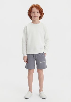 Παιδί με κόκκινα μαλλιά που στέκεται, φοράει λευκό πουκάμισο με μακριά μανίκια, γκρι σορτς με πλαϊνές τσέπες και λευκά παπούτσια χωρίς κορδόνια, σε απλό φόντο.