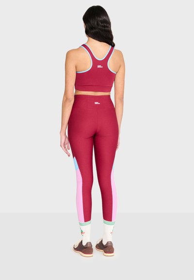 Femme aux longs cheveux foncés, vue de dos, portant un brassard de sport bordeaux et des leggings avec des panneaux roses et bleus, des chaussettes blanches et des baskets marron.
