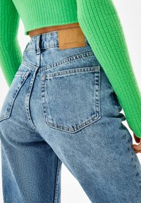 Vaaleansiniset farkut korkealla vyötäröllä, joissa on kaksi takataskua ja nahkalappu. Kirkasvihreä ribbiknitti-pusero näkyy ylhäällä.