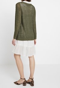 Maglione lavorato a maglia di colore verde oliva con trama aperta, abbinato a un vestito bianco a strati con pois e scarpe con stampa leopardata.