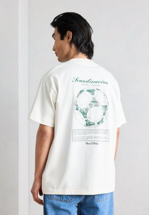 Junger Mann mit schwarzen Haaren, der ein weißes T-Shirt mit einem grünen Vintage-Fußballmotiv und dem Schriftzug „Scandinavian Football Community“ auf dem Rücken trägt.