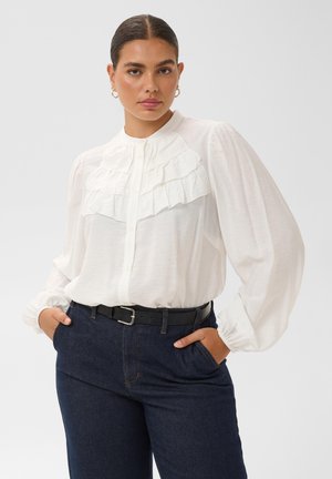 Jeune femme aux cheveux foncés attachés porte une blouse blanche à volants, un jean bleu foncé et une ceinture noire, posant avec les mains dans les poches.
