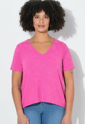 Kobieta o kręconych włosach, ubrana w jaskraworóżowy top z krótkim rękawem i dekoltem w serek oraz niebieskie dżinsy, stojąca na białym tle.