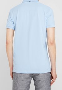 Polo bleu clair à manches courtes avec col. Fabriqué en tissu doux, présentant une texture lisse et un ourlet droit. Un pantalon gris est visible.