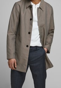 Man i brun rutig jacka över vit zip-neck tröja och mörkgrå byxor med en hand i fickan mot en grå bakgrund.