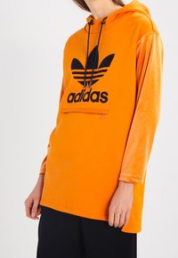 Person som bär en orange Adidas-hoodie med svart logotyp, långa ärmar, framficka med dragkedja och huva med snörning, över svarta byxor.