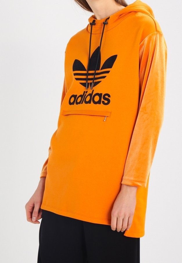 Person som bär en orange Adidas-hoodie med svart logotyp, långa ärmar, framficka med dragkedja och huva med snörning, över svarta byxor.