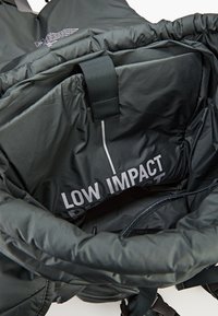 Schwarzes, gepolstertes Innenfutter des Rucksacks mit glänzender Oberfläche, das ein großes, in Weiß gedrucktes Etikett mit der Aufschrift "LOW IMPACT" und verstellbaren Trägern aufweist.
