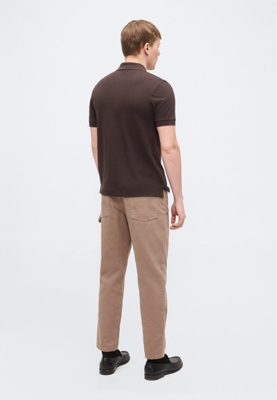 Homme debout de dos, portant un polo à manches courtes marron foncé, un pantalon beige, des chaussettes noires et des chaussures noires sur un fond uni.