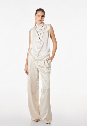 Vrouw draagt mouwloze ivoorkleurige satijnen top en wijde satijnen broek, geaccentueerd met lange ketting en gouden creolen, staat met hand in zak.