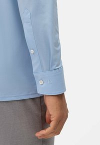 Lichtblauwe langarmshirt met knoopmanchetten. Heeft een gladde stof, een taillefit en decoratieve stiksels nabij de manchette.