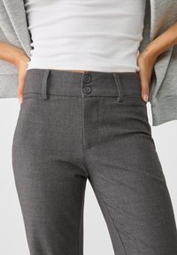 Pantalons gris sur mesure avec une texture lisse, dotés de deux boutons noirs à la taille, d'une coupe ajustée et de poches latérales pour plus de fonctionnalité.