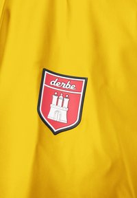 Giacca impermeabile gialla con una patch a forma di scudo rosso con un design a torre e la scritta "derbe" in bianco. Tessuto liscio e leggero.