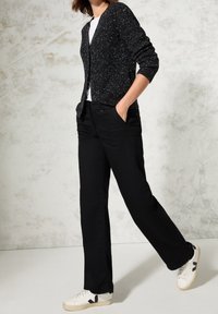 Czarny dziergany kardigan z białymi kropkami, biała koszulka i czarne spodnie, połączone z białymi trampkami z czarnymi akcentami, pozycja stojąca.